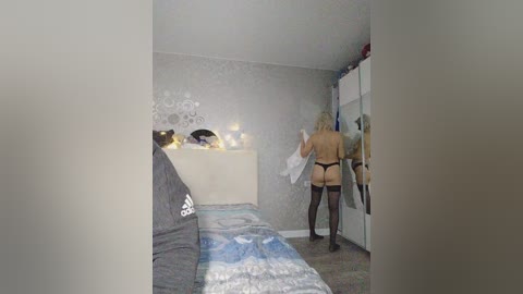 A video of a bedroom with a blonde woman in black lingerie and stockings, facing away, standing near a bed with grey bedding and a white headboard. The room has a grey carpet and silver wallpaper.