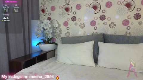 Media: Video of a modern bedroom with a white bed, gray headboard, and a floral-patterned wallpaper. A white orchid plant sits on a dark wooden nightstand. A digital screen shows a user's Instagram post.