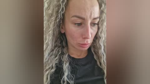 Video of a woman with curly, silver-grey hair and fair skin, wearing a black top, looking introspective. The background is plain, beige walls.
