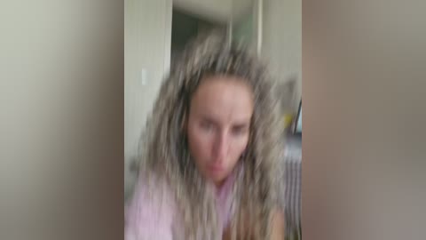 A blurry video of a woman with long, curly blonde hair wearing a light pink shirt, taken indoors. The background features a beige wall and indistinct furniture.