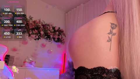 Video of a fair-skinned woman in a black lace bra, with a tattoo of a rose on her right shoulder, in a dimly lit room with pink and purple lighting.