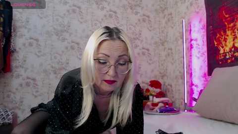 Media: A blonde woman with glasses and red lipstick leans forward, wearing a black sheer top, in a room with floral wallpaper, a bed, and a pink lit fireplace.