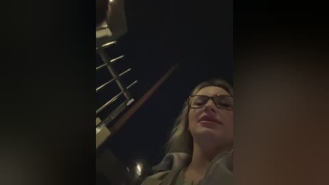 Video of a woman with glasses and light brown hair, wearing a grey top, sitting in a dimly lit room with a staircase and railing in the background.