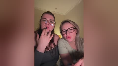 Video of two women kissing, one with glasses and long hair, the other with blonde hair and glasses, in a dimly lit room with beige walls.
