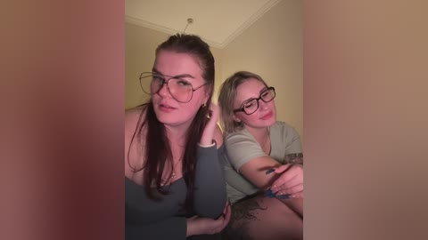 Video of two young women with fair skin, wearing glasses and dark clothing, sitting closely together on a bed, partially obscured by a person standing in front.