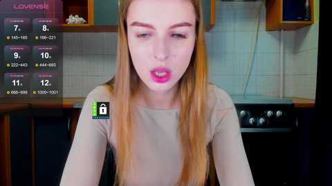 A video of a fair-skinned woman with long blonde hair, wearing a beige top, making a kissing gesture in a modern kitchen with wooden cabinets and white tiles.