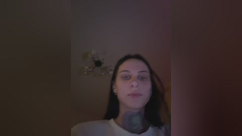 Video of a young woman with long, dark hair, pale skin, and a neutral expression, standing in a dimly lit room with a beige wall and a small, white flower arrangement.