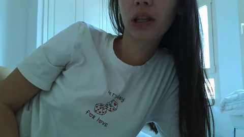 Video of a young woman with long, straight brown hair, wearing a white t-shirt with \"I roll for love\" and dice print. Background shows a white room with a bed and window.