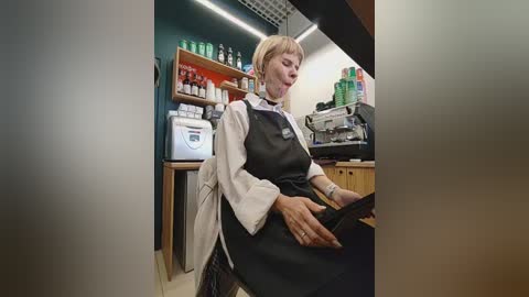 Video of a white, middle-aged woman with short blonde hair in a salon, wearing a white lab coat over a black apron, working on a customer's hair.