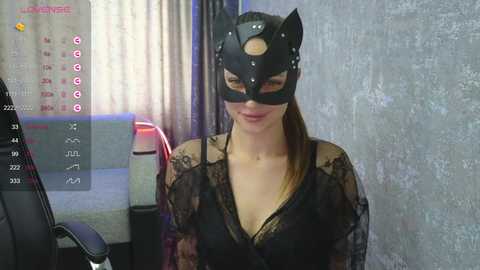 A video of a woman in a black lace dress and cat mask, standing in a dimly lit room with a calendar on the wall and a desk.