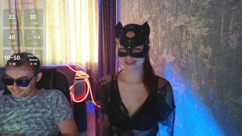 Video of a man in a gray T-shirt, sunglasses, and a woman in a black lace dress with a cat mask, sitting in a dimly lit, graffiti-covered room.