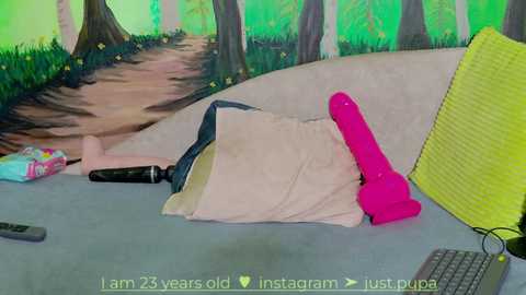 Media: Video of a young woman with pink hair, lying on a gray floor, surrounded by a vibrant green forest mural. She is nude, with a pink vibrator and a pink dildo nearby.