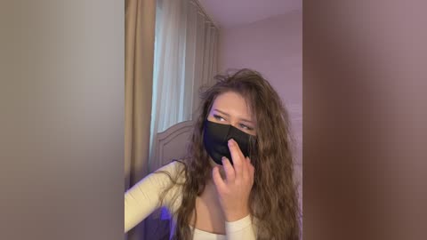 Video of a young woman with long, wavy brown hair, wearing a black face mask, white long-sleeve top, and beige curtains in the background, standing in a dimly lit room.