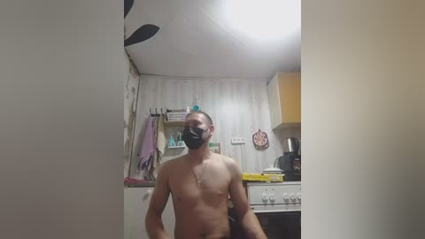 Media: Video of a shirtless, muscular man wearing a black face mask, standing in a cluttered kitchen with a white wall, hanging towels, and a stove in the background.
