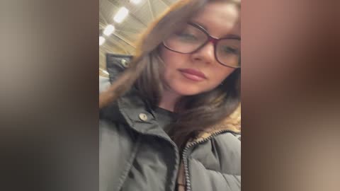 Media: Video of a young woman with fair skin, brown hair, and glasses, wearing a gray puffer jacket, standing in a dimly lit, blurred indoor setting.