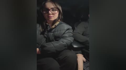 Media: Video of a woman with glasses and dark hair in a black puffy jacket, sitting in a dimly lit car, looking introspective.