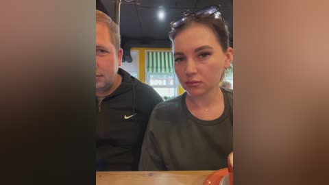 Media: Video of a blonde man and brunette woman seated at a wooden table in a dimly lit, modern restaurant. They wear casual attire; man in black jacket, woman in gray top.