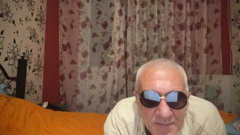 Media: Video of an elderly man with short white hair, wearing sunglasses and a beige shirt, sitting on a bed with orange sheets. Background features floral-patterned curtains and wallpaper.