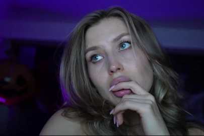 Media: Video of a young woman with light skin, long blonde hair, and blue eyes, looking thoughtful with her hand to her mouth, set against a dimly lit, blurred background with purple lighting.