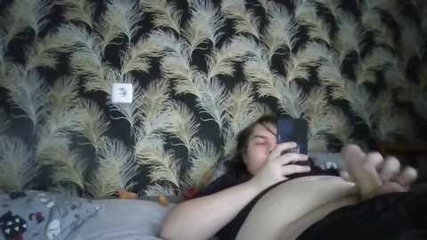 Video of a plus-sized woman with light skin, dark hair, and black clothing, lying on a bed with gray sheets. She holds a smartphone, capturing a selfie. Background features a wallpaper with an abstract pine tree pattern.