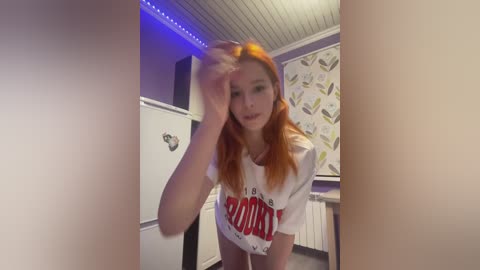 Video of a young woman with long, straight red hair, wearing a white \"Hooters\" t-shirt, bending over to adjust her glasses in a dimly lit room with a patterned wall and blue LED lighting.