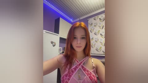 Video of a young woman with long, straight red hair, wearing a pink floral-patterned bralette, sitting in a room with purple walls, white ceiling, and a leaf-patterned wall hanging.