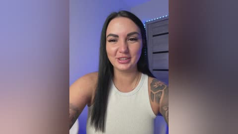 A video of a light-skinned woman with long black hair and tattoos on her arms, smiling, wearing a white tank top, in a dimly lit room with blue lighting.