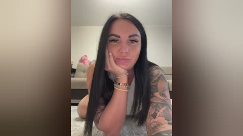 Video of a tattooed, dark-haired woman with long hair, wearing a gray top, making a pouty face on a carpeted floor.