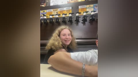 Media: Video of a smiling young woman with curly blonde hair, wearing a black shirt and white arm cast, sitting in a fast food restaurant.