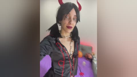 Media: Video of a person in a black, red-ribboned, and horned devil cosplay outfit with black hair, red lipstick, and a silver chain necklace, standing in a purple and red room.