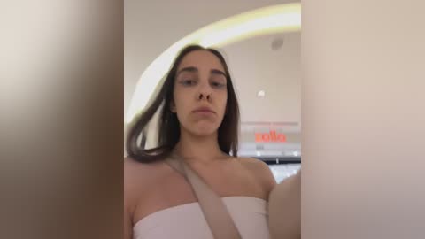 Media: A video of a young woman with long dark hair, fair skin, and a neutral expression, wearing a strapless white top, seated in a car with a beige seatbelt. The background features a blurred interior with an arched window and a \"Dolce & Gabbana\" sign.