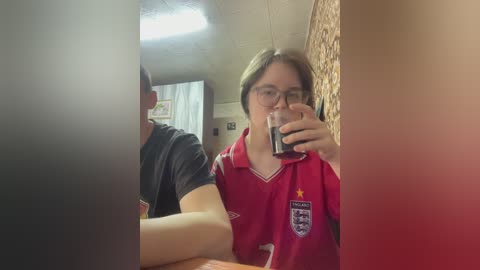 Video of a young woman with short brown hair, wearing glasses and a red England football shirt, taking a selfie in a bathroom.