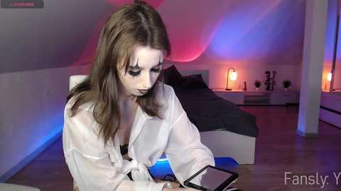 Media: Video of a young woman with fair skin and long brown hair, wearing a sheer white top, focusing on her smartphone in a modern bedroom with purple and blue lighting, and a bed with black sheets.