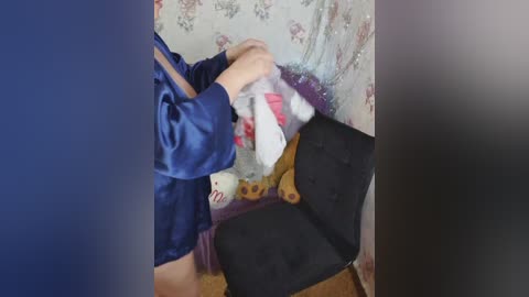 A video shows a person with light skin, wearing a blue jacket and holding a white and red blanket, sitting on a black chair against a floral-patterned wall.