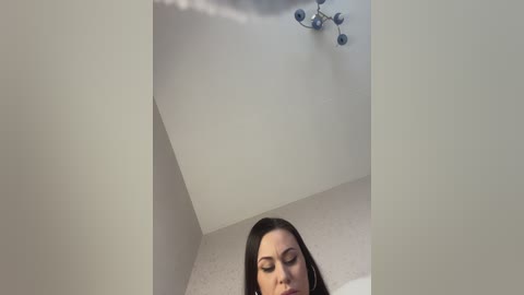 Video of a woman with long, dark hair and closed eyes, standing in a white-walled room with a ceiling light fixture.