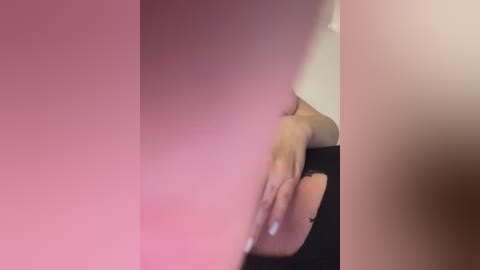 A blurry video with a pinkish tint features a person's hand, partially obscured by a large, dark object. The hand, with a simple black bracelet, is seen holding something. The background is indistinct.