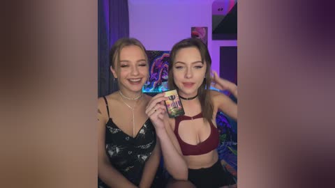 Media: Video of two young women with fair skin and brown hair, smiling, wearing black and red lingerie, sitting on a colorful bed with a tapestry background.