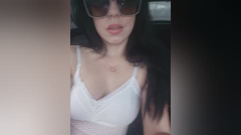 Media: Video of a young woman with long dark hair, wearing large sunglasses, a white lace top, and sitting in a car.