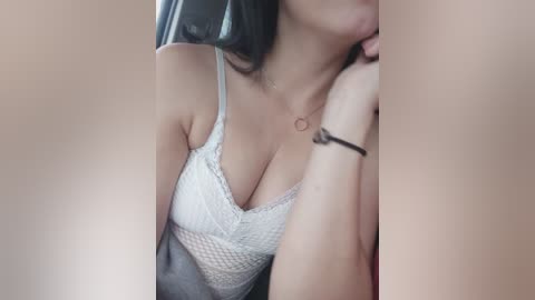 A close-up video of a light-skinned woman with shoulder-length black hair, wearing a white lace bralette that accentuates her ample cleavage. She is resting her chin on her hand, and her arm is adorned with a black bracelet. The background is blurred.