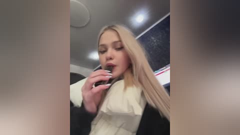 Media: Video of a young blonde woman with fair skin, wearing a black jacket and beige top, applying lipstick in a dimly lit room with a starry ceiling.