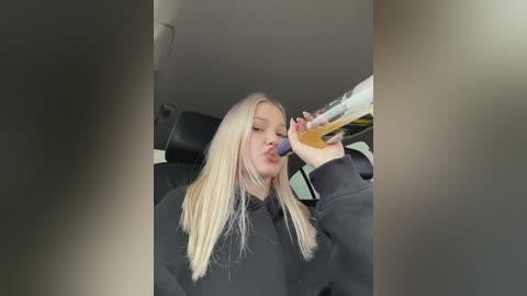 Media: Video of a young blonde woman drinking beer from a bottle while sitting in the passenger seat of a car, wearing a black hoodie.