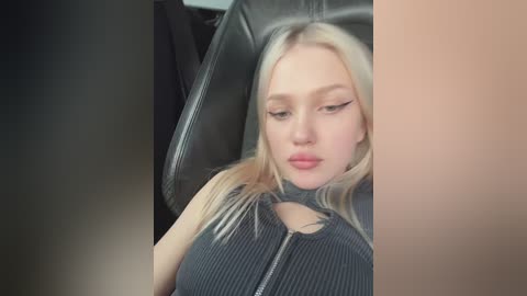 Media: Video of a blonde woman with fair skin and full lips, wearing a dark gray, ribbed crop top, sitting in a black leather car seat.