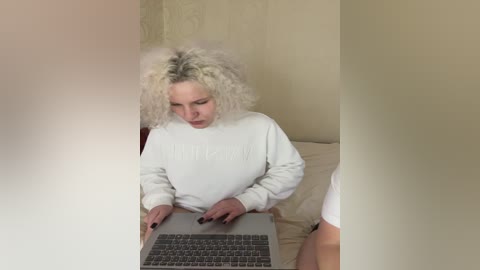 Media: A video of a curly-haired woman in a white sweatshirt, typing on a silver laptop, sitting on a bed with beige walls and white bedding.