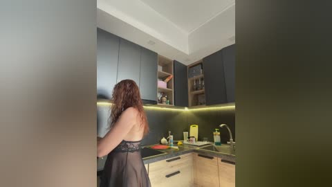 Video of a woman with long red hair, wearing a sheer black lace dress, standing in a modern kitchen with dark cabinets, stainless steel appliances, and a yellow countertop.
