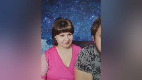 Video of a smiling, light-skinned woman with shoulder-length brown hair in a bright pink top, standing beside a man with short brown hair, against a starry space-themed backdrop.