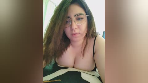 Video of a fair-skinned woman with long brown hair and glasses, wearing a black camisole that reveals ample cleavage, taken indoors with blurred background.