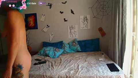 A video of a woman in a room with gray walls, adorned with various drawings and spider webs. She stands nude, facing the camera, with a blue bedspread and pillows in the background.