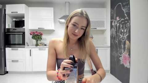 Media: Video of a blonde woman in a modern kitchen, holding a can of shaving cream, wearing a strapless gray top. White cabinets, black stove, and chalkboard wall with a floral drawing.