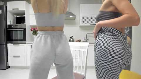 Media: Video of two women in a modern kitchen. One wears a gray crop top and baggy sweatpants, the other a black crop top and black-and-white checkered leggings.