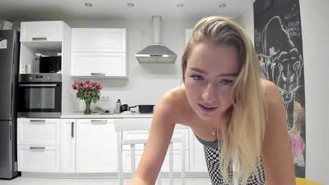 Media: Video of a blonde woman with long hair in a black-and-white checkered dress, leaning over a kitchen counter. White cabinets, blackboard wall, and stainless steel appliances in a modern kitchen.